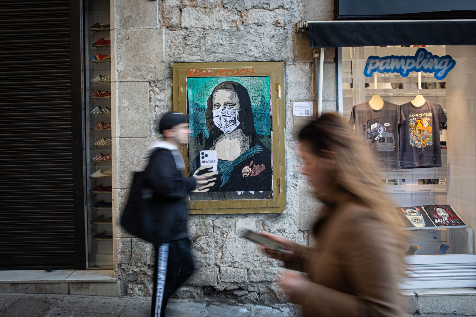 Dos personas pasan con sus móviles junto a una pintura de una Gioconda con un móvil y una mascarilla, obra del grafitero TV Boy llamada 'Mobile World Virus', en referencia a la cancelación del Mobi