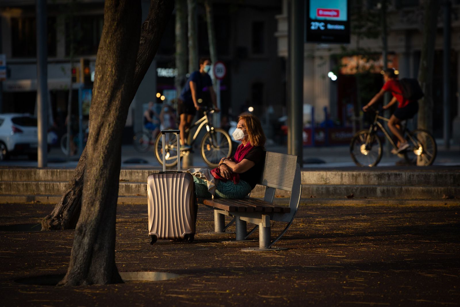 Una mujer sentada en un banco junto a una maleta, mientras varios ciclistas pasan por detrás, en Barcelona, Catalunya (España), a 28 de julio de 2020.