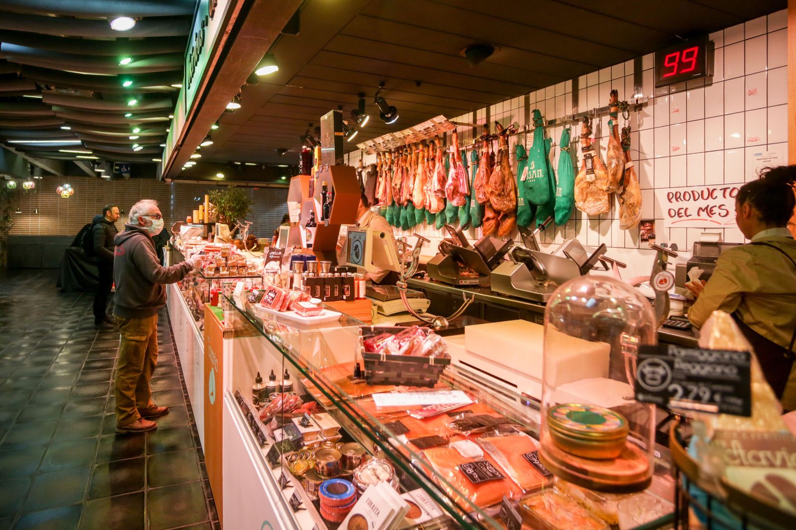Varios clientes hacen la compra en un puesto de carne y embutidos en el mercado de San Antón, que permanece abierto y anuncia que también atiende a domicilio, en Madrid (España), a 18 de marzo de 2