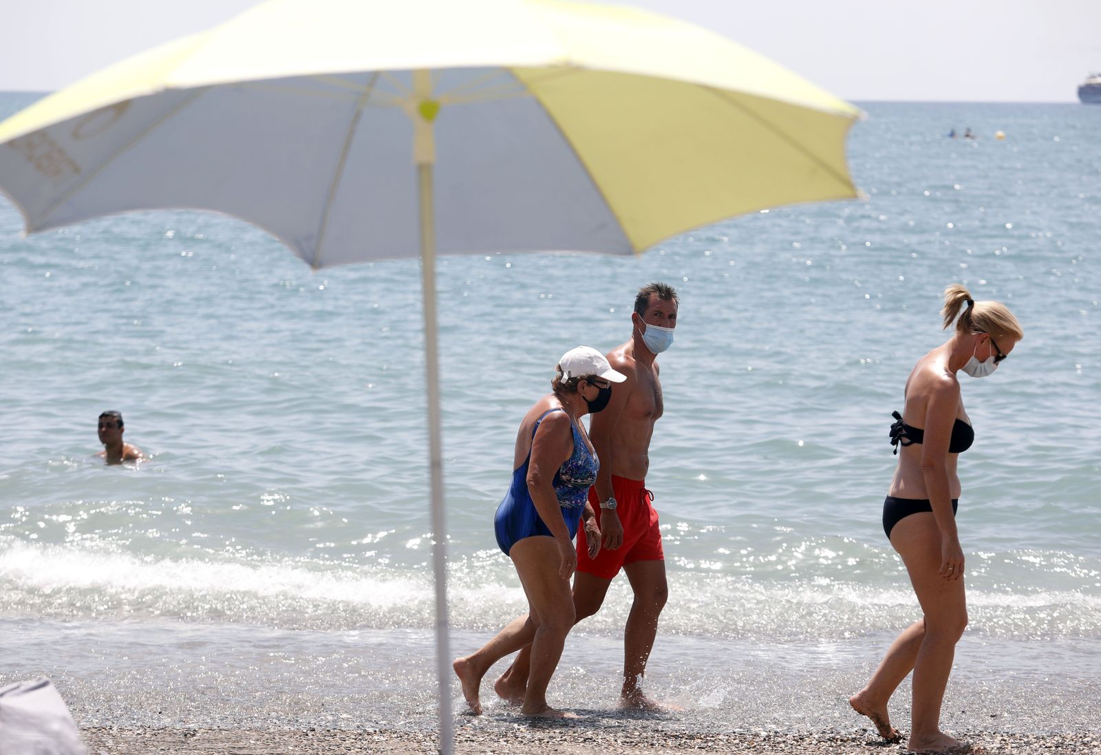 Bañistas con mascarilla pasean por la orilla de la playa.